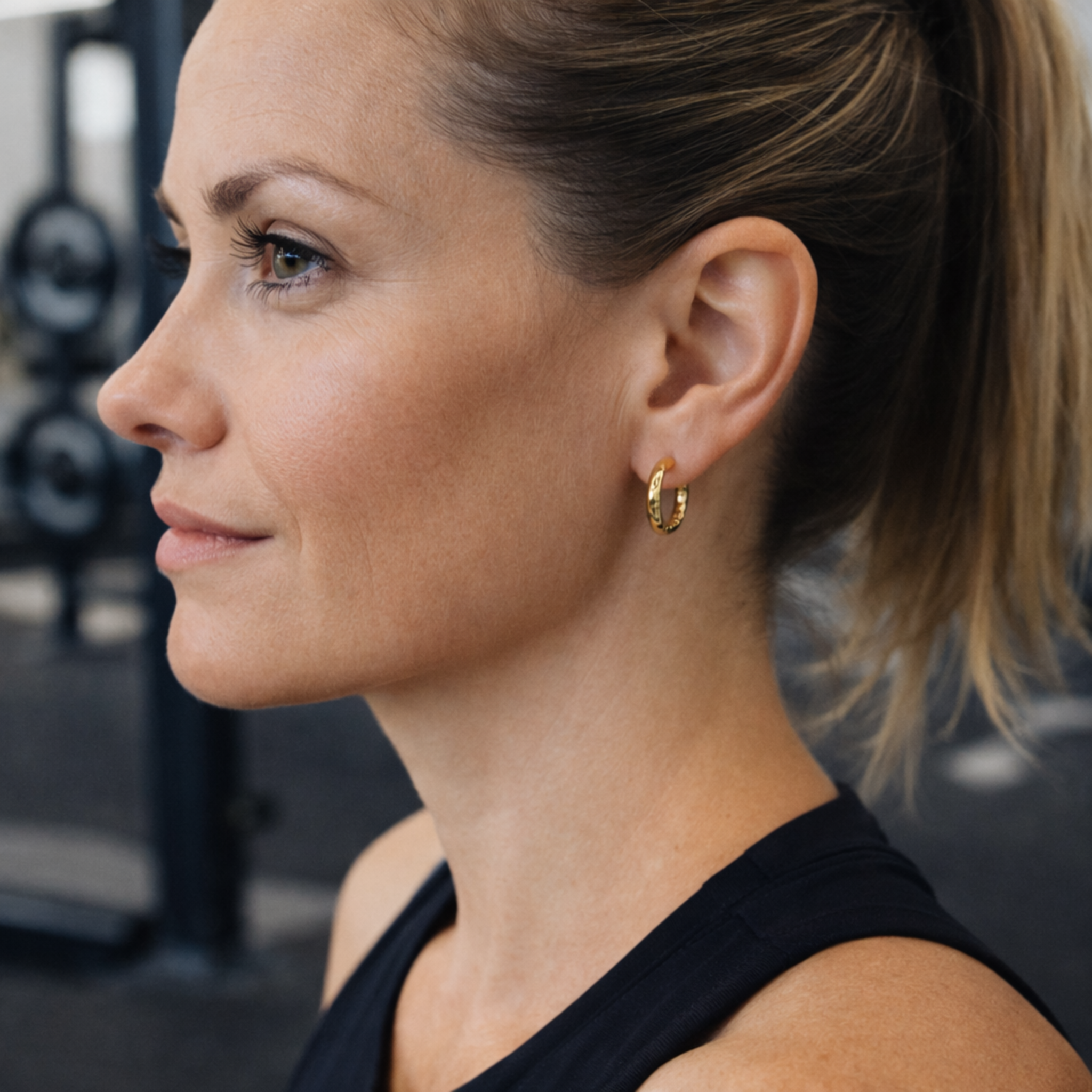 Female model wearing gold 12mm Aura hoop earrings with a high polish 18K gold PVD finish, shown in a close-fitting lightweight everyday style at the gym
