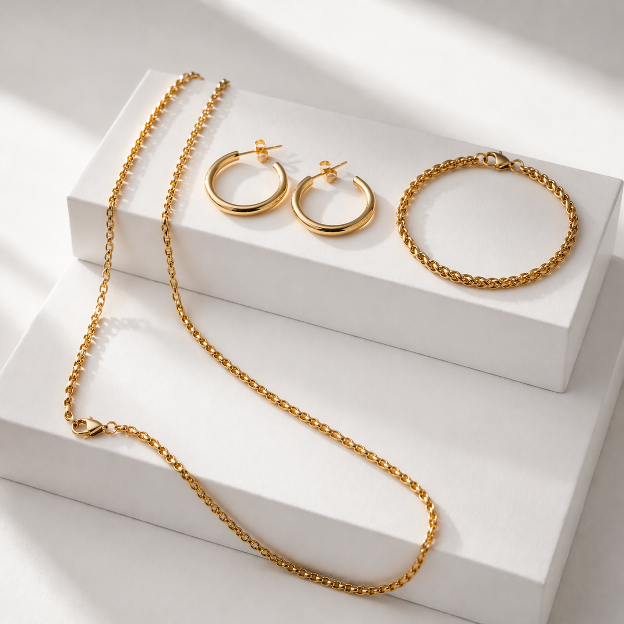 Essential Set jewelry featuring 18K gold PVD waterproof necklace, bracelet and hoop earrings displayed on clean white blocks, tarnish resistant gold jewelry set
